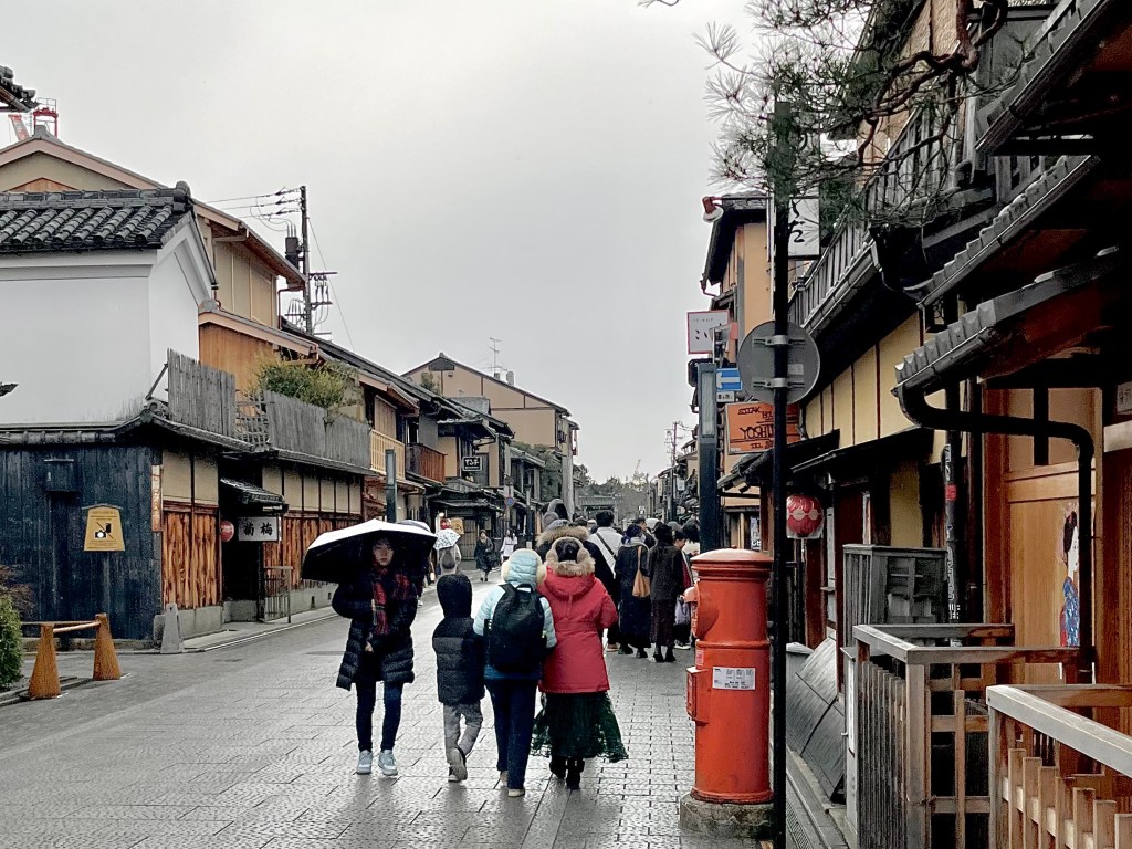 雨天的京都祇園花見小路，街道兩旁是傳統日式町家建築，遊客撐傘行走在石板路上，右側可見復古紅色郵筒與藝妓插畫，充滿懷舊和風情懷。 A rainy day on Hanamikoji Street in Kyoto’s Gion district, lined with traditional wooden machiya houses. Visitors walk along the stone-paved street under umbrellas. A classic red postbox and a geisha illustration can be seen, evoking a nostalgic atmosphere.