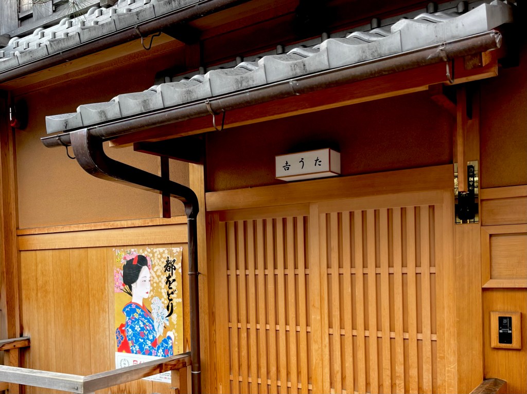 京都祇園花見小路的傳統木造茶屋門口，木格拉門旁張貼著一幅穿著華麗和服的藝妓海報，店名招牌為「吉うた」，屋簷覆蓋著灰色瓦片，充滿日式風情。 Entrance of a traditional wooden teahouse on Hanamikoji Street in Kyoto's Gion district. A poster of a geisha in a colorful kimono is displayed beside the sliding wooden door. The sign above reads "Yoshiuta", and the tiled roof completes the classic Japanese aesthetic.