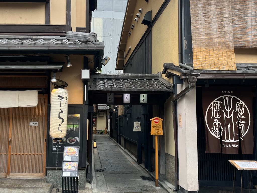 京都祇園花見小路旁的小巷入口，傳統町家建築環繞，左側懸掛著寫有「きらら」的白色日式燈籠，右側為掛布簾的老店鋪，石板小路延伸至巷弄深處，氣氛靜謐充滿和風情懷。 An entrance to a narrow side alley off Hanamikoji Street in Kyoto’s Gion district. Traditional wooden machiya buildings line both sides. A white lantern with the word “Kirara” hangs on the left, while a classic noren curtain marks a historic shop on the right. The stone-paved path leads into a quiet, nostalgic passageway.