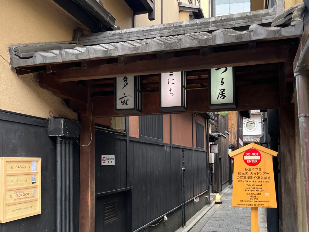 京都祇園花見小路某處巷弄入口，設有木造日式屋簷與店家燈籠招牌，下方設有「禁止進入」與「私人道路」的告示牌，提醒遊客禁止拍照與擅入，尊重當地居民生活空間。 A narrow alleyway entrance off Hanamikoji Street in Kyoto's Gion district, marked with traditional lantern signs and a wooden roof. A bold “DO NOT ENTER” sign warns tourists not to enter or take photos, as it is a private road.