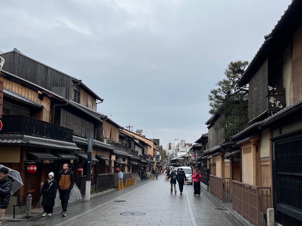 京都祇園花見小路的街景，兩側為整齊排列的木造町家建築，街道鋪設石板，部分遊客撐傘或散步於街上，展現日式古都的寧靜氛圍。 A wide view of Hanamikoji Street in Kyoto’s Gion district, lined with preserved wooden machiya buildings. The stone-paved street is filled with strolling visitors, capturing the serene charm of Japan’s traditional townscape.