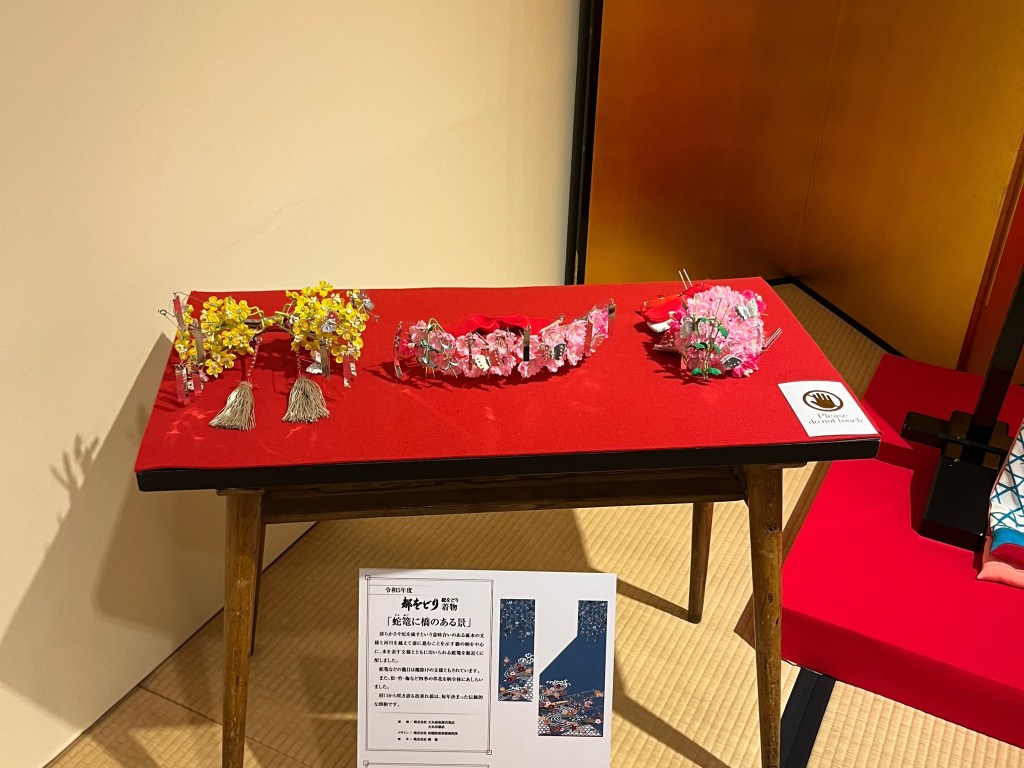 京都祇園花街藝術資料館中展示的舞妓髮飾，包括以金屬線與絲花製作的季節性簪花，擺放於紅布展示台上。