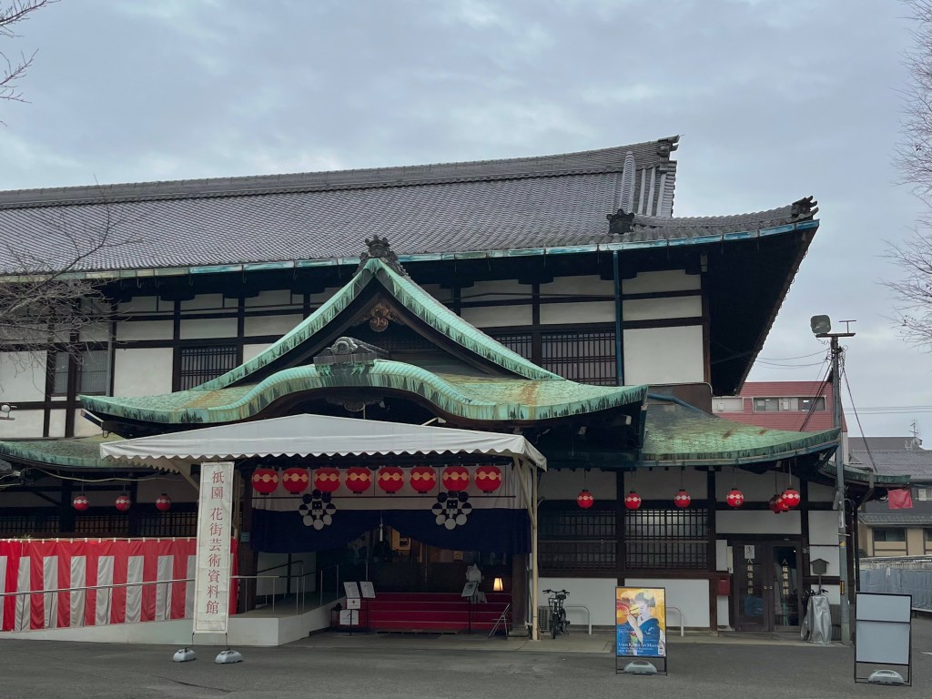 京都祇園花街的傳統建築－花街藝能資料館外觀，屋簷綠銅瓦與日式格窗展現古典風格，門前掛有紅色燈籠與祭典布幕。
