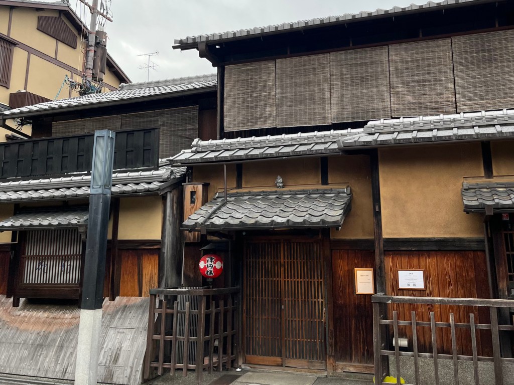 京都祇園花見小路的傳統茶屋外觀，木造牆面與格子門展現和風美感，屋簷上方掛有竹簾與紅底白紋的圓形燈籠，為藝伎活動的代表標誌。 A traditional teahouse facade on Hanamikoji Street in Kyoto's Gion district. The wooden exterior, sliding lattice door, bamboo blinds, and iconic red lantern evoke the classic atmosphere of geisha culture.