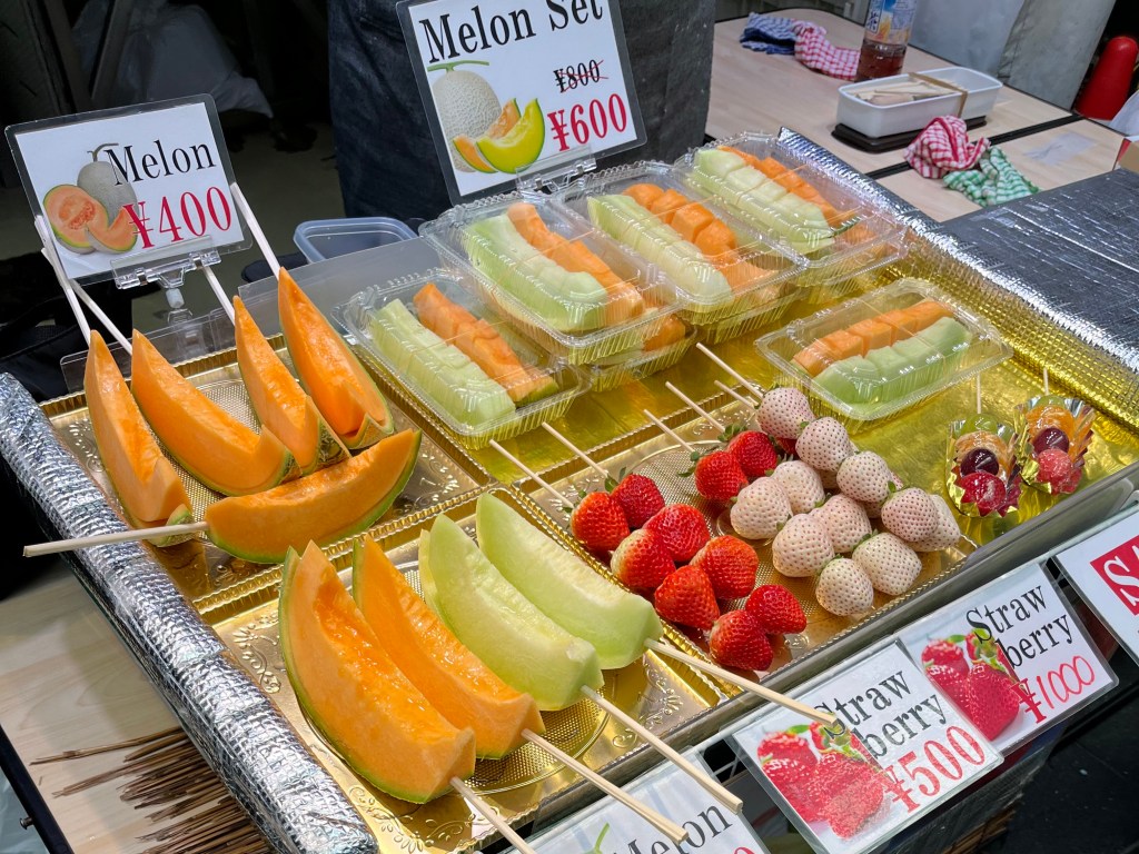 黑門市場販售各式水果的攤位，展示哈密瓜、綠肉香瓜、草莓、白草莓與水果串，標示日圓售價。