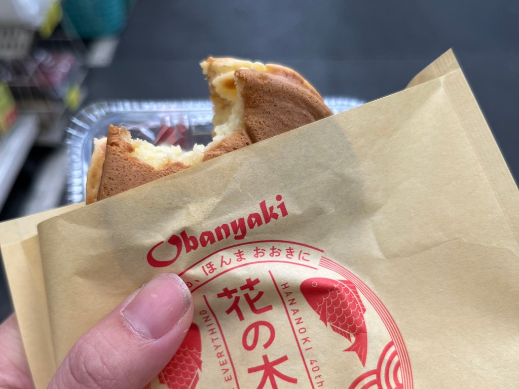手持剛咬一口的大判燒（Obanyaki），包裝袋印有「花の木」與鯛魚圖案，背景為黑門市場街道。
