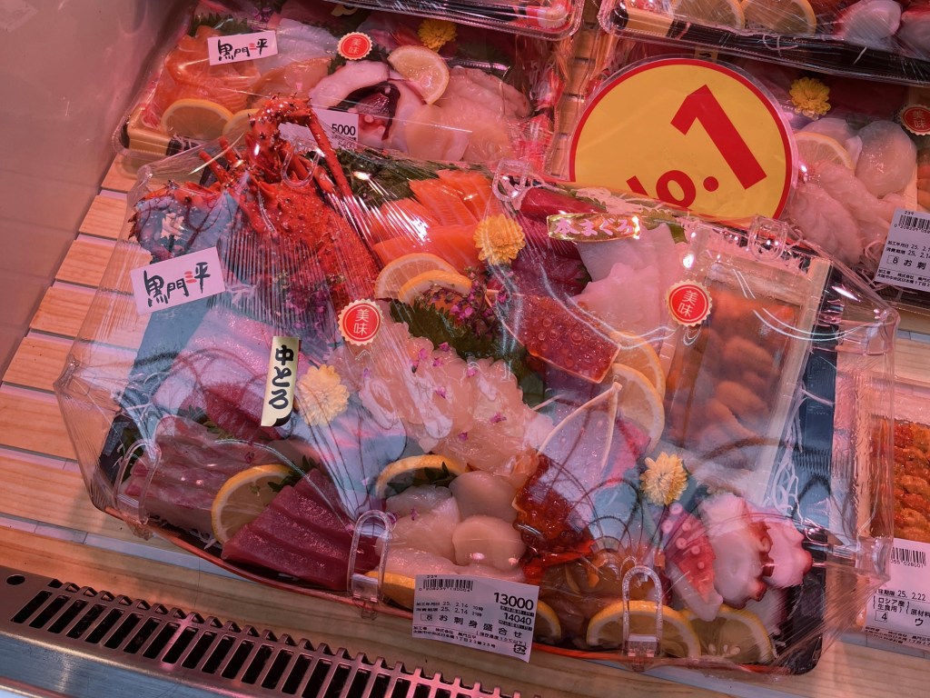黑門市場販售的豪華生魚片拼盤，包含中腹鮪魚、鮭魚、章魚與蝦，裝於透明盒中，價格標示為13000日圓。