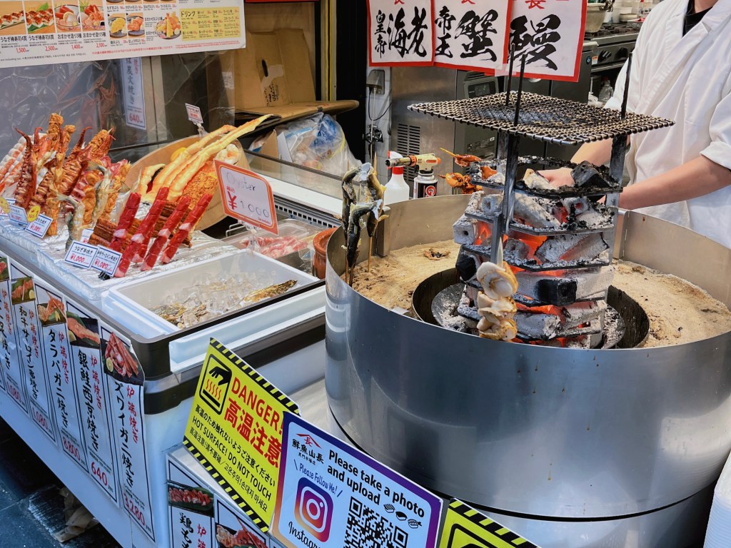 黑門市場攤販使用炭火現烤海鮮串燒，包括香魚與扇貝，旁邊設有高溫警示標語。